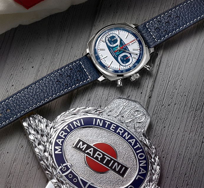 {generated} Breitling Top Time Martini Racing watch with blue strap shown beside a polished Martini racing emblem on wood